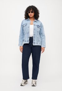 Chaqueta de denim azul claro con cierre de botones y bolsillos, llevada sobre una blusa blanca. Combinada con jeans azul oscuro y zapatillas con estampado.