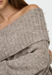 Pull surdimensionné en tricot de couleur brun clair avec une texture côtelée et un design décolleté. Présente des manches longues et un tissu épais et doux.