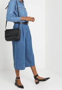 Koszula dżinsowa, niebieskie kuloty, czarna torebka o teksturowanym wykończeniu oraz czarne baleriny na szpilce z paskiem wokół kostki i zapięciem.