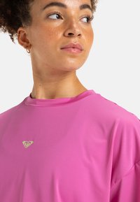 Camiseta deportiva de color rosa brillante, transpirable, con mangas cortas, que presenta un pequeño logo verde en el pecho y una tela suave y ligera.