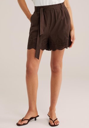 Braune Shorts mit hoher Taille, gewelltem, besticktem Saum und gebundenem Stoffgürtel, getragen mit braunen Flip-Flop-Sandalen mit Absatz.