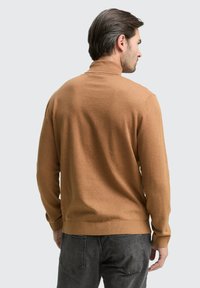 Maglione marrone a maniche lunghe con collo alto, polsini e orlo a costine. Indossato sopra jeans grigio scuro. Texture morbida, design minimale.