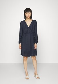 Vestido envolvente azul-marinho com mangas bufantes longas, tecido texturizado, decote em V e cintura ajustada, caindo logo acima do joelho.