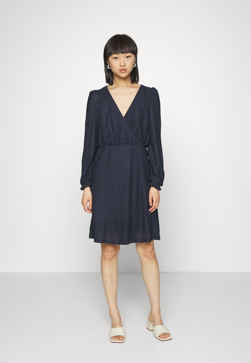 Vestido envolvente azul-marinho com mangas bufantes longas, tecido texturizado, decote em V e cintura ajustada, caindo logo acima do joelho.