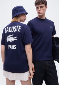 Δύο μοντέλα φορούν μπλε ναυτικού χρώματος μπλουζάκια Lacoste· το ένα δείχνει την πίσω πλευρά με το λογότυπο "LACOSTE PARIS" και το κροκόδειλο, ενώ το άλλο κοιτάει μπροστά κρατώντας μια τσάντα.
