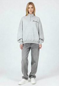 Sweat-shirt en polaire grise avec col haut et fermeture éclair au quart, orné d'une broderie noire. Associé à un jean en denim gris clair et des baskets blanches.