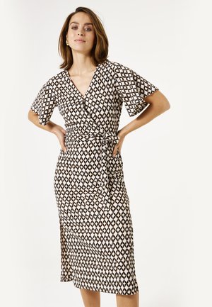 Creme kleurige wikkeljurk met een geometrisch zwart en bruin patroon, korte mouwen en een strikceintuur, met een lengte tot onder de knie.