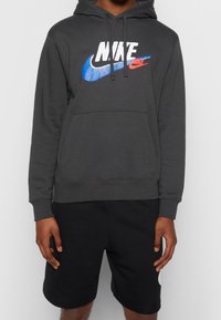 Šedá mikina vyrobená z bavlněné směsi, s modrým a červeným logem Nike potištěným přes hrudník a přední klokaní kapsou. Nosená s černými šortkami.