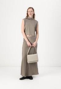 Helle beige ärmellose Bluse mit hohem Kragen, kombiniert mit einem passenden langen Rock. Das Model hält eine hellgraue Handtasche und trägt schwarze Schuhe.