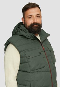 Grüne Puffweste mit Kapuze, ausgestattet mit schwarzen Druckknopftaschen, rotem Reißverschluss und gestepptem Design, getragen über einem hellen Pullover.