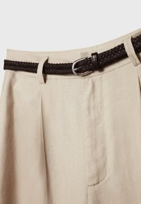 Gros plan sur un pantalon beige avec des plis et une ceinture tressée brun foncé passée dans les passants, sécurisée par une boucle argentée.