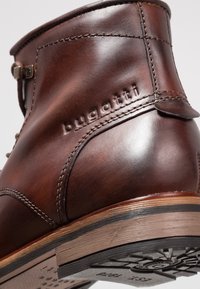 Brązowy skórzany botek sięgający do kostki, z fakturowanym wykończeniem, detalami przeszyć i niewielkim obcasem. Wytłoczone logo z boku. Gumowa podeszwa z wzorem.