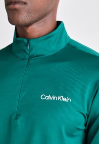 Pullover verde con zip e tessuto testurizzato. Presenta un colletto alto e un logo bianco "Calvin Klein" sul lato sinistro del petto.