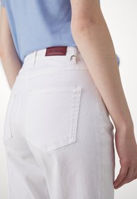 Pantalons en denim blanc avec une coupe ajustée, des poches arrière et une étiquette de marque en cuir bordeaux près de la taille. Texture lisse et lignes épurées.