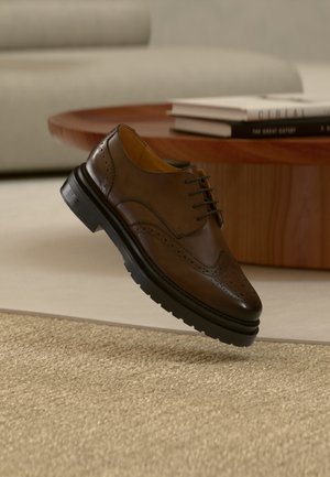 Brauner Leder-Brogue-Schuh mit schwarzer Sohle schwebt über einem beigen Teppich, im Hintergrund ein Holztisch und gestapelte Bücher.
