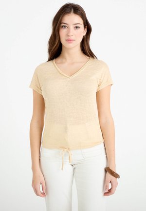 Ung kvinde med langt brunt hår iført beige kortærmet V-hals top og hvide bukser, stående foran en ensfarvet hvid baggrund.