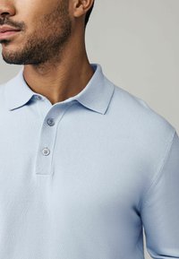 Hellblaues Polohemd mit Kragen, drei Knöpfen und strukturiertem Stoff. Nahaufnahme des Oberkörpers.