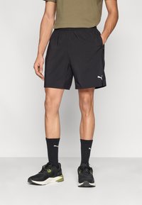 Μαύρο αθλητικό σορτς με ελαστική μέση και πλαϊνές τσέπες. Φορεμένο με μαύρα αθλητικά παπούτσια με πράσινες λεπτομέρειες και μαύρες κάλτσες.