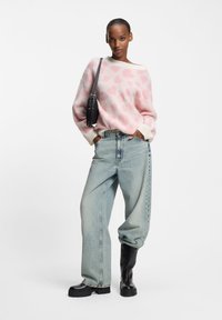 Jasnoróżowy sweter z wzorami serc, kremowe mankiety, do tego luźne, jasnoniebieskie dżinsy i czarne botki na platformie. Czarna torba na ramieniu.