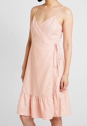 Femme portant une robe portefeuille rose pâle sans manches avec des bretelles fines et un ourlet à volants, nouée à la taille, sur un fond uni.