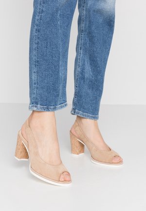 Beige suede open-toe heels with a cork block heel, featuring a white sole and a cut-out design. Worn with blue rolled denim jeans.