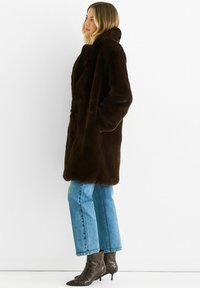 Brown faux fur coat with a high collar, featuring a button closure and two side pockets. Paired with blue cropped jeans and patterned boots.