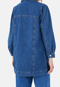 Farkkutakki, jossa on rento istuvuus, pitkät pussimaiset hihat ja napitettu hihansuut. Tummansininen väri, näkyvillä olevilla saumoilla ja takana olkasauman yksityiskohdilla.