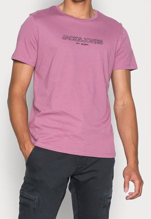 Mężczyzna w gładkim różowym t-shircie z napisem „JACK&JONES EST. MCMXC”, w czarnych spodniach typu cargo, z rękami swobodnie opuszczonymi wzdłuż ciała.