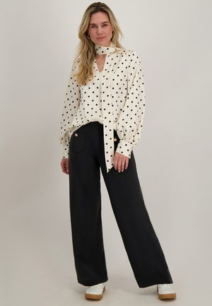 Vrouw die een witte polkadot blouse met een nekstrik draagt, zwarte wijde broekspijpen en witte sneakers, staand tegen een effen achtergrond.