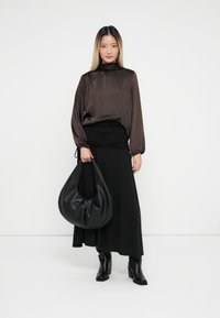 Woman wearing a dark brown satin blouse and black skirt, holding a large black leather handbag, standing against a white background.