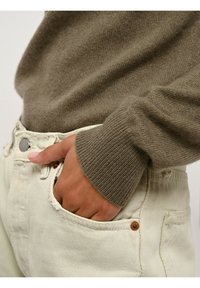 Brauner Strickpullover mit gerippten Bündchen, getragen über weißen Denim-Jeans mit sichtbarem Knopf und Tasche. Weicher, texturierter Stoff.