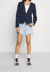 Blazer marine avec un bouton en or, associé à une jupe en denim clair avec un ourlet effiloché et une fente. Baskets blanches avec des accents colorés.