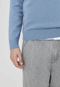 Un pull en maille bleu clair avec un ourlet et des poignets côtelés, associé à un pantalon texturé gris clair. Une main repose sur le côté.