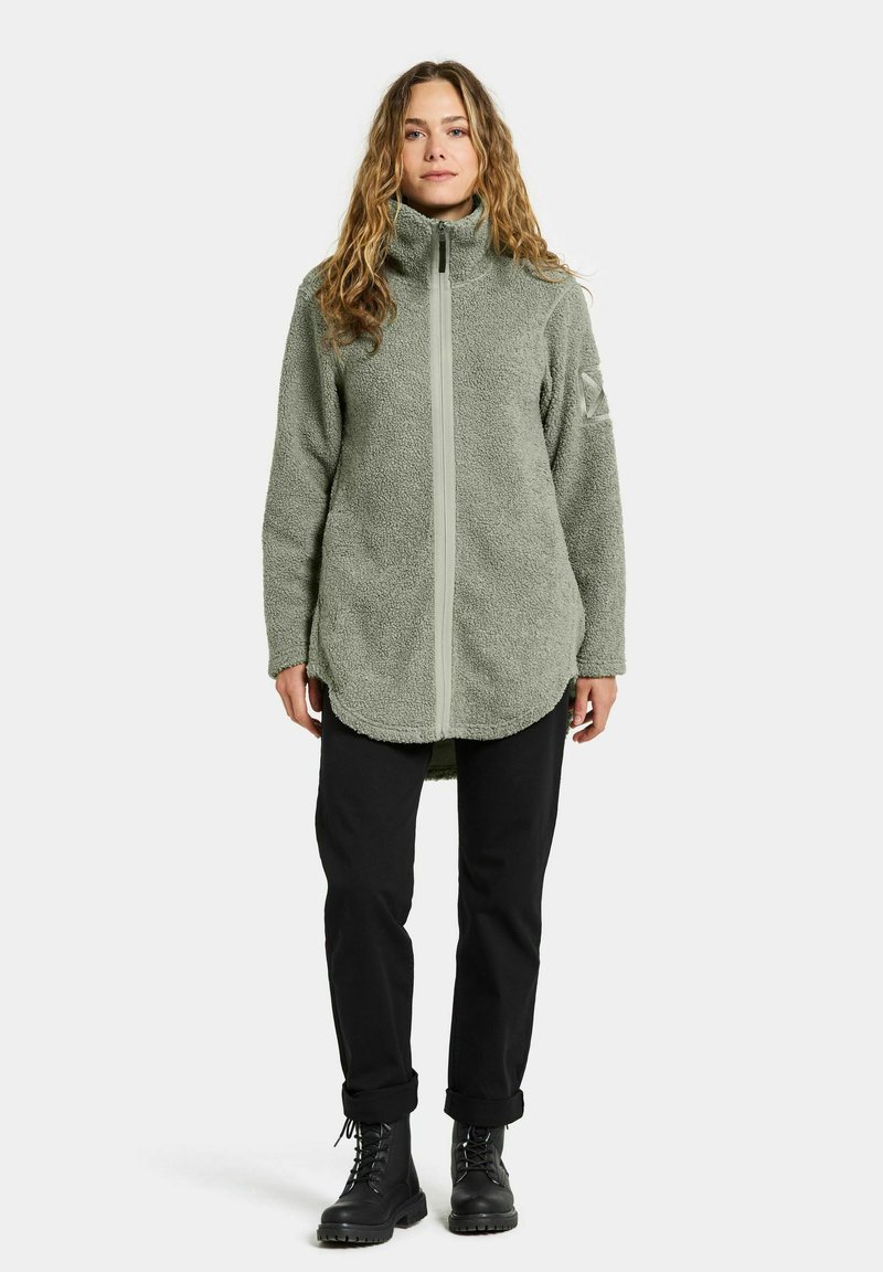 Fleecejacke in Hellgrün, mit hohem Kragen, vollem Frontreißverschluss und seitlichen Taschen. Kombiniert mit schwarzen Hosen und Schnürstiefeln.