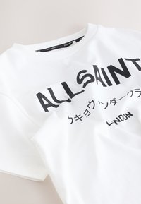 T-shirt blanc AllSaints avec texte anglais et japonais en noir et une étiquette de marque noire à l'intérieur du col.