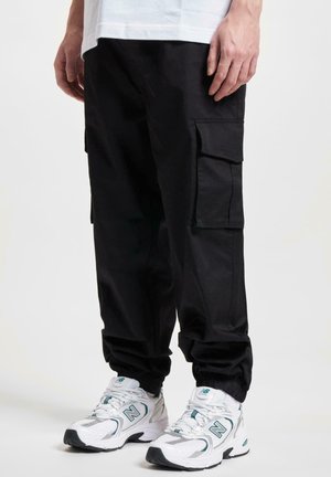 Pantaloni cargo negri cu multiple buzunare laterale, manșete elastice și o textură netedă, asortați cu adidași albi cu accente turcoaz.