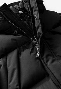 Svart puffjacka med en texturerad insida, med en framdragkedja och logotyp dragflik. Platta sömmar och vadderade sektioner synliga.