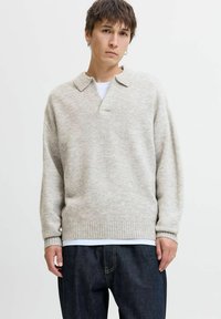 Lys grå strikket sweater med polo-krave og ribbede manchetter og kant. Båret over en hvid t-shirt og parret med mørke denimbukser.