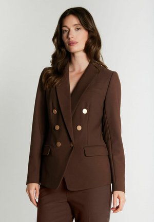Femme aux longs cheveux bruns portant un blazer croisé marron foncé sur mesure et un pantalon assorti, debout devant un fond uni.