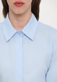 Camicia azzurra chiara con colletto, dotata di una fila di bottoni e una texture liscia. Presenta una leggera lucentezza e cuciture rifinite con cura.