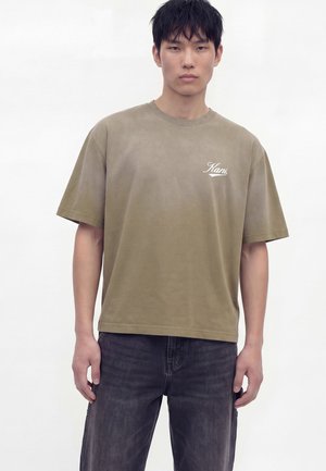 Mladý muž s rovnými čiernymi vlasmi má na sebe oversized khaki tričko s logom "Kani" a vyblednuté čierne rifle, stojí pred jednofarebným pozadím.