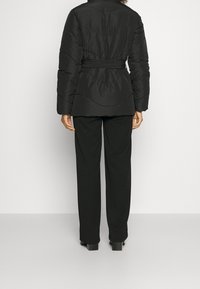 Giubbotto nero imbottito con vita con cintura, caratterizzato da motivi trapuntati testurizzati. Indossato sopra pantaloni neri abbinati, mostrando una silhouette elegante.