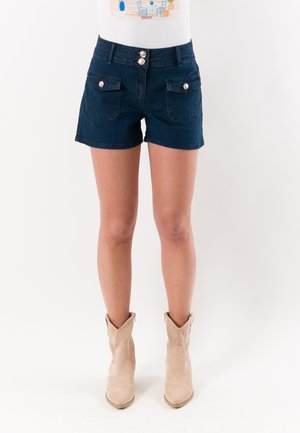 Gambe che indossano pantaloncini di denim blu scuro con tasche anteriori a patta e stivaletti beige in suede su uno sfondo bianco semplice.