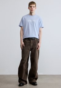 Svetlomodré bavlnené tričko s bielym nápisom „Calvin Klein“, skombinované s voľnými tmavohnedými nohavicami a čiernym opaskom, na jednoduchom pozadí.