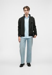 Ung mann står iført lys blå skjorte, svart jakke, lys blå jeans, svarte sko, og bærer en svart skulderveske foran en hvit bakgrunn.