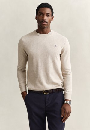 Mann mit Schnurrbart, der einen beigen Pullover, marineblaue Hosen, einen geflochtenen Gürtel und eine silberne Armbanduhr trägt und mit den Händen in den Taschen steht.