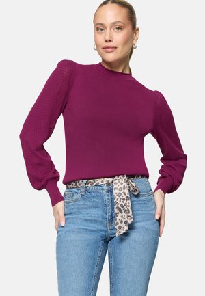 Femme portant un pull moulant violet avec des manches bouffantes texturées et un jean taille haute bleu noué avec une ceinture imprimé léopard.