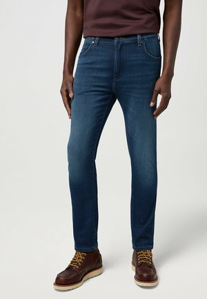 Dark blue denim jeans with a slim fit, featuring five pockets and subtle fading. Worn with brown boots and a maroon t-shirt.