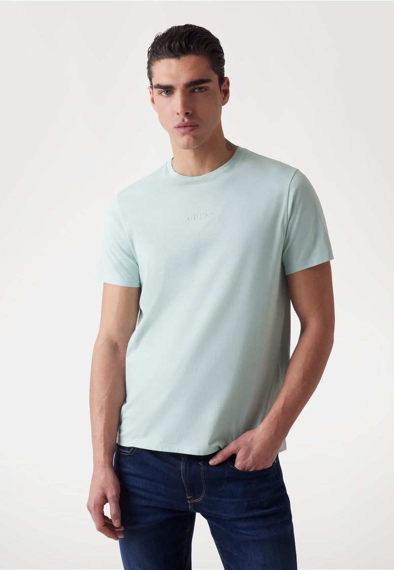 Lichtblauwe shirt met korte mouwen en ronde halslijn, met een reliëf "GUESS" logo op de borst, gecombineerd met donkerblauwe jeans.