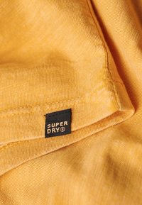 Tessuto giallo con una consistenza liscia, caratterizzato da un'etichetta nera con la scritta "SUPER DRY" in lettere dorate. Visibili dettagli delle cuciture irregolari.
